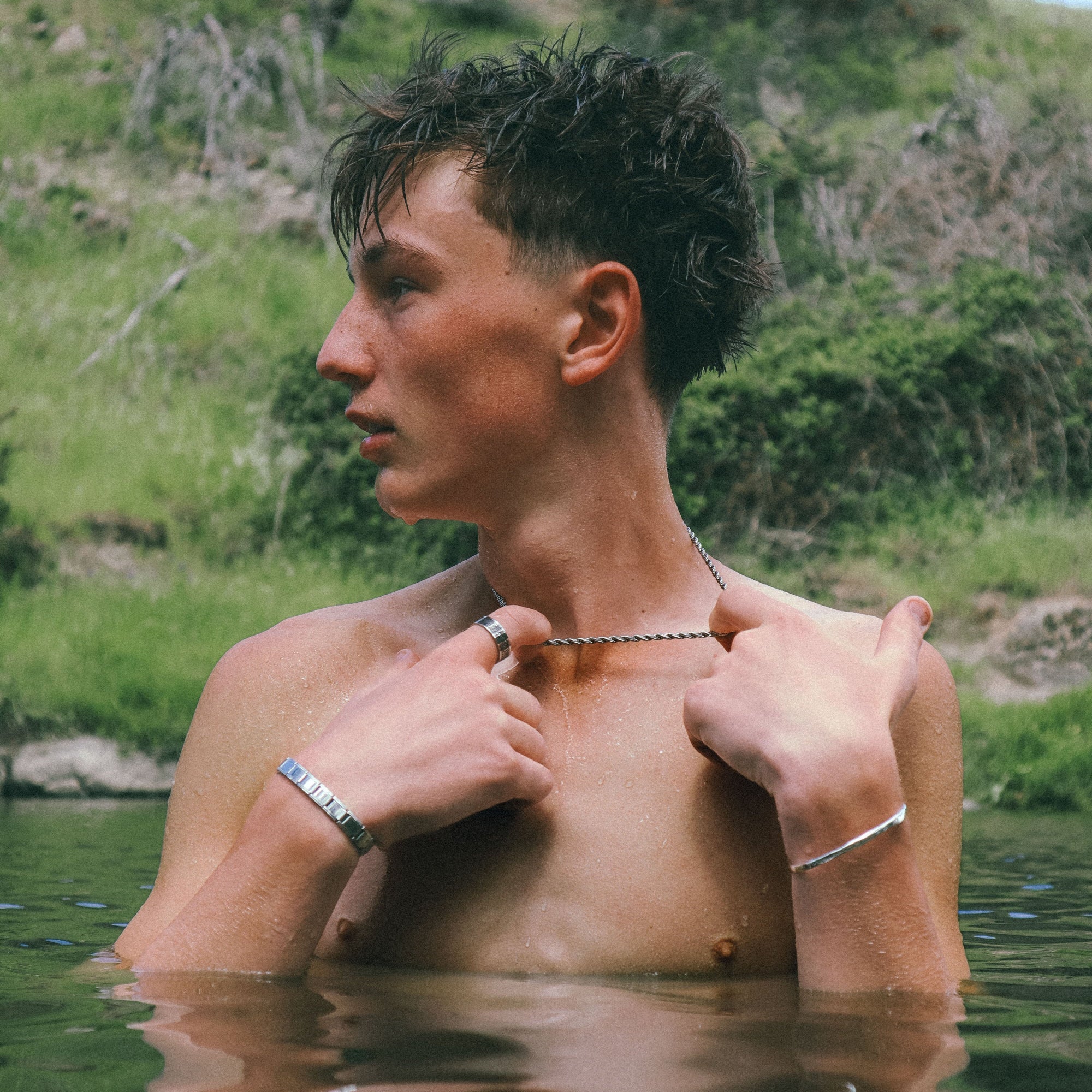 Man adjusting a necklace by a body of water with a reflective surface.