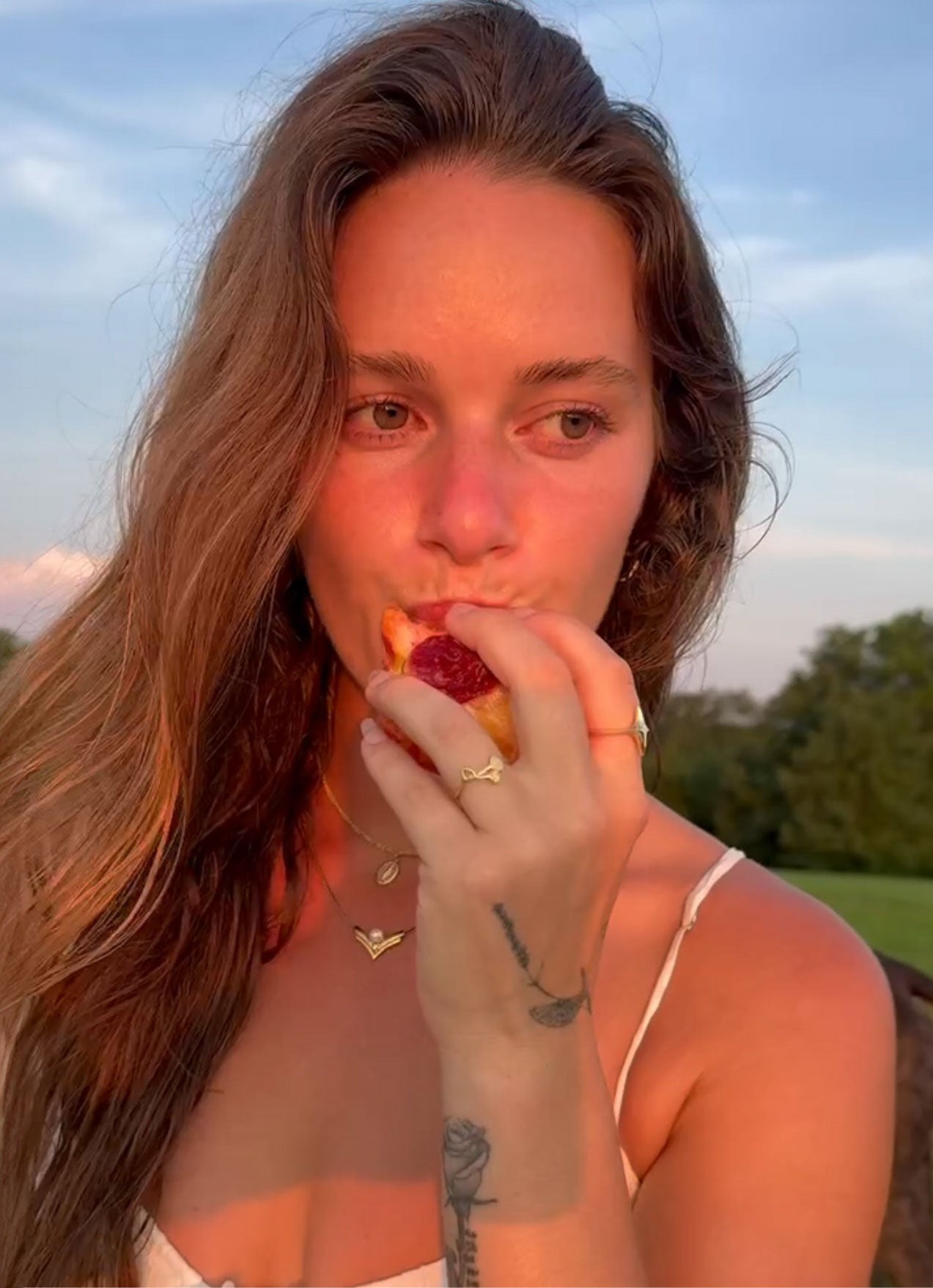Woman eating a pastry outdoors with a scenic background. showing gold ginkgo ring on hand.