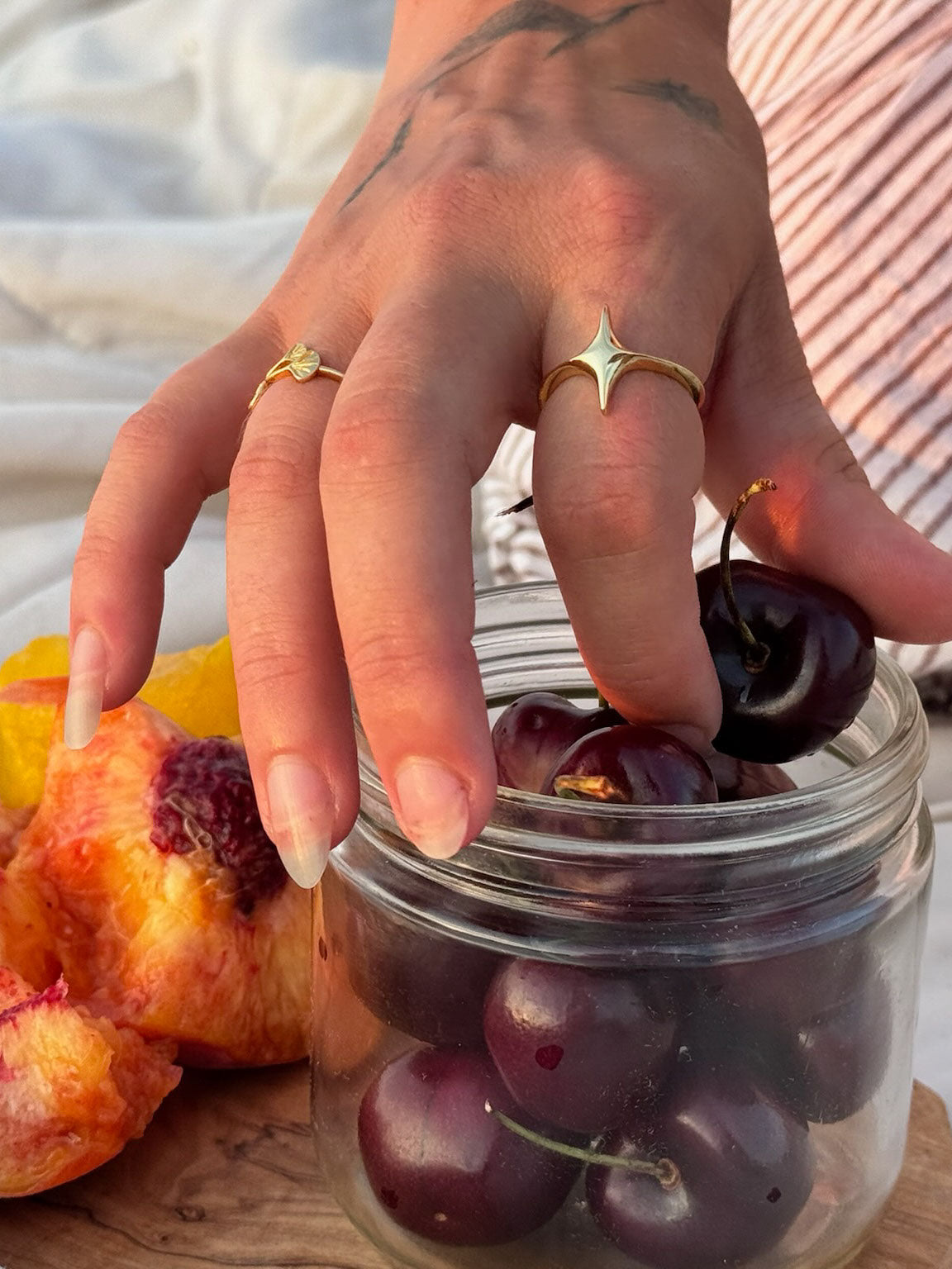 Hand with gold rings picking cherries from a jar on a wooden surface, showing star ring and ginkgo leaf ring