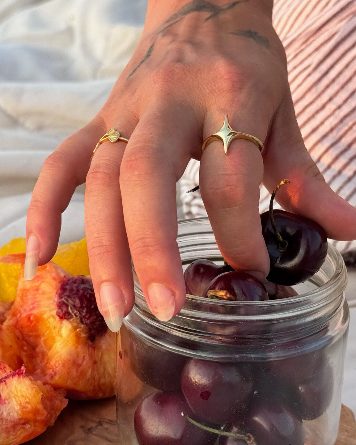 Hand with gold rings picking cherries from a jar on a wooden surface, showing star ring and ginkgo leaf ring
