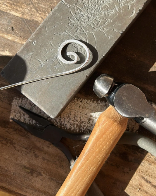 Metalworking tool with a spiral design on metal, surrounded by tools on a wooden surface.
