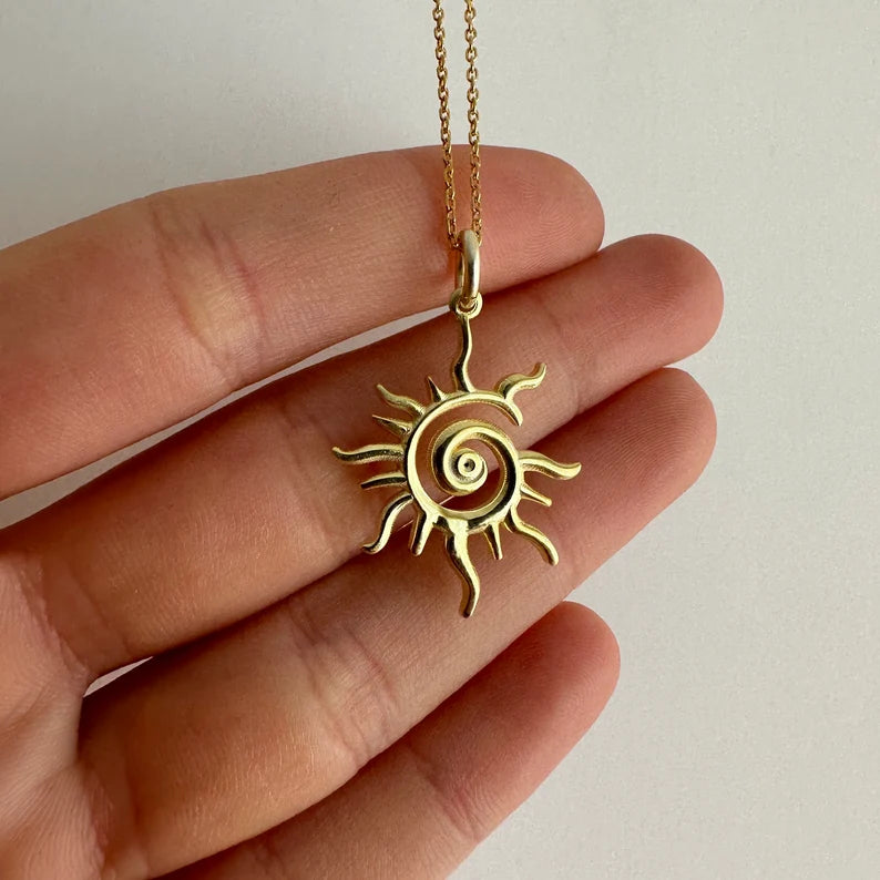 Gold sun-shaped pendant on a chain held between fingers against a neutral background