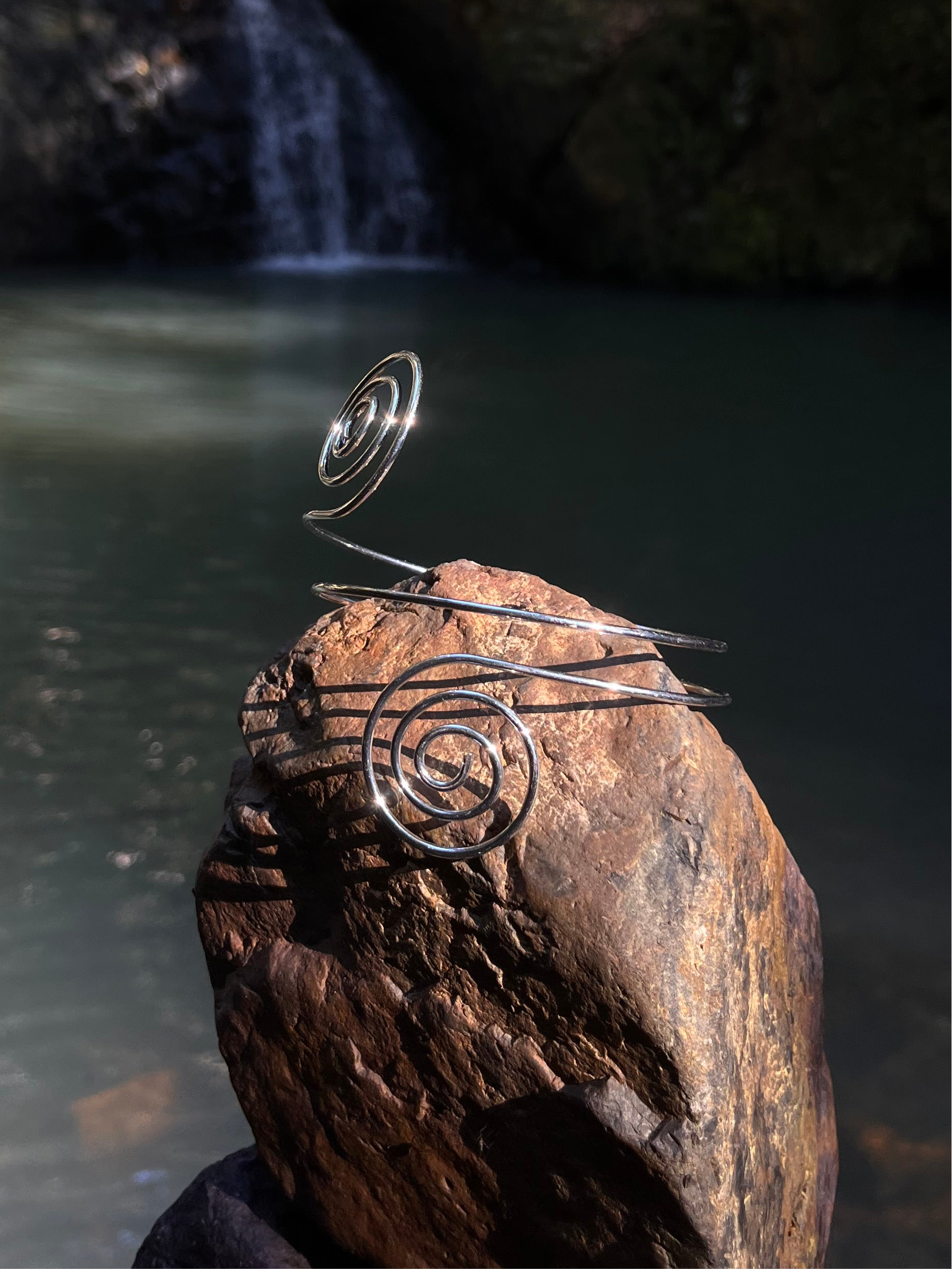 A silver cuff bracelet with a spiral design displayed on a rock by a waterfall.
