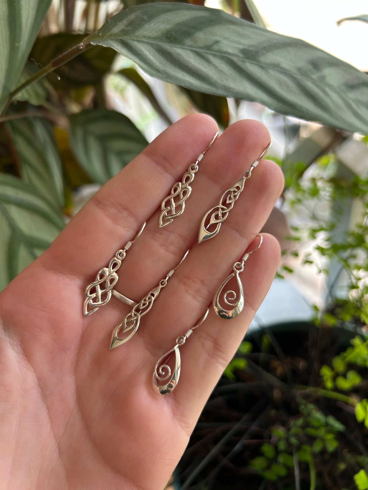 Hand holding multiple Yallambe handmade recycled silver drop earrings with delicate cut out designs beside indoor plants.