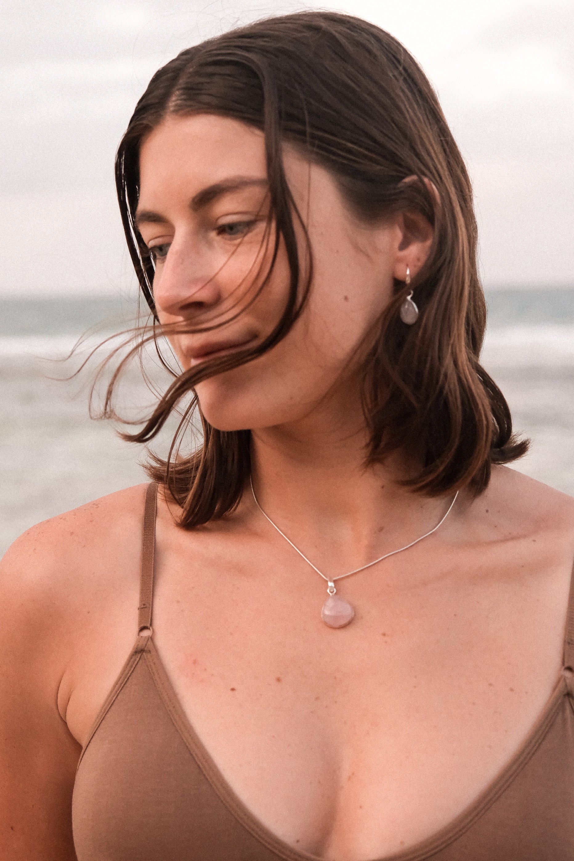 A person wearing a brown tank top and showcasing a silver pendant necklace with a rose quartz charm, standing near a beach.