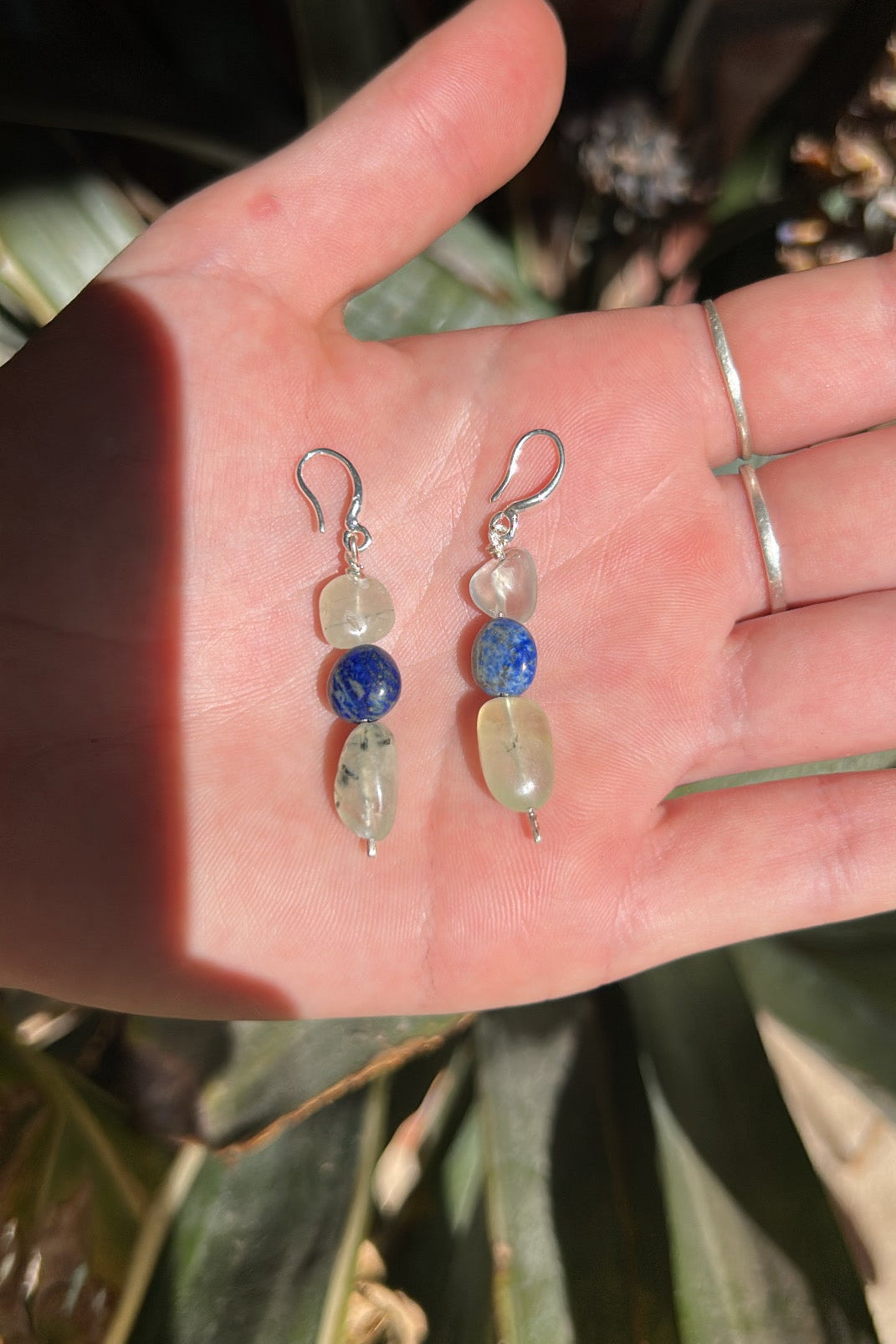 Hand holding two beaded earrings with a plant in the background