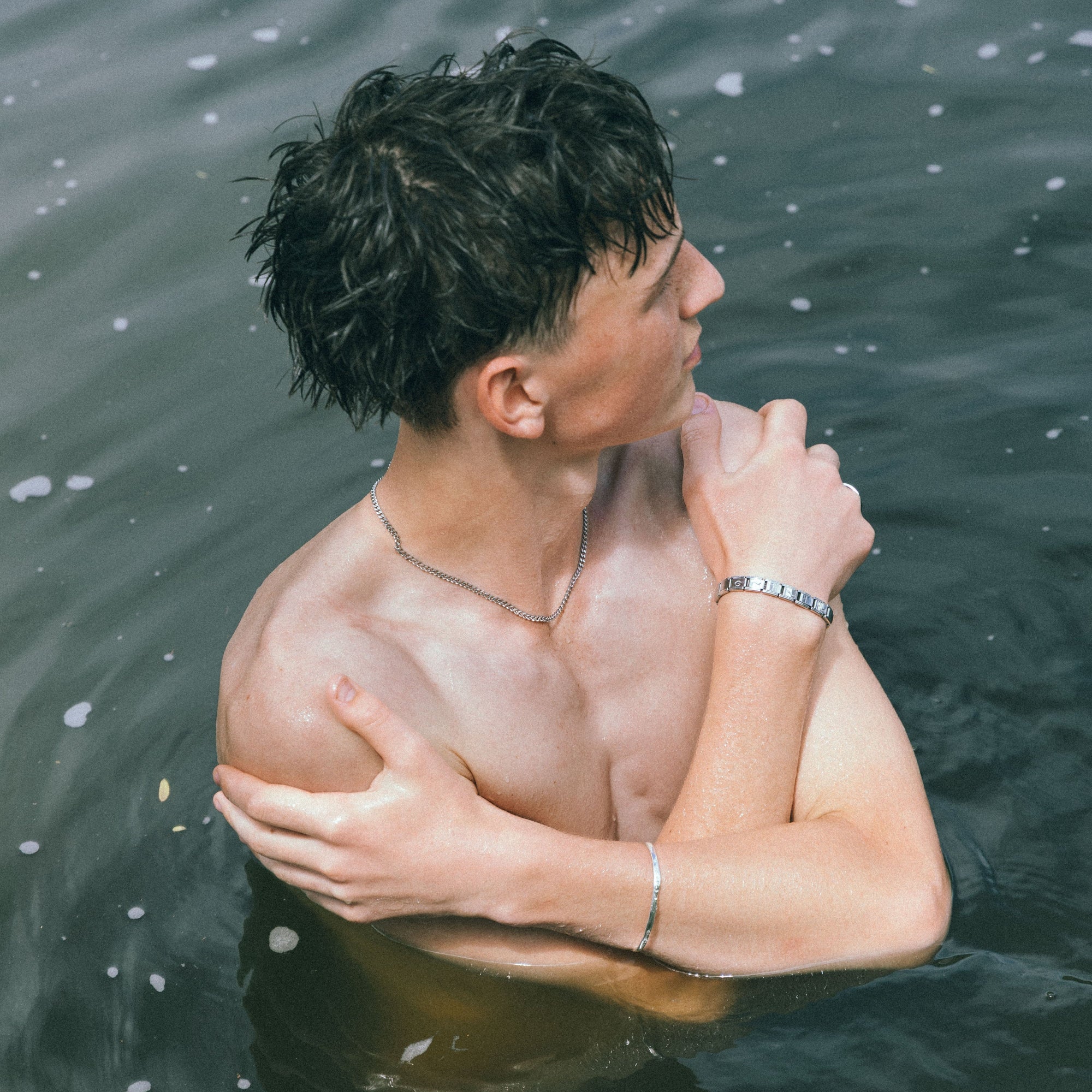 Man with a muscular build wearing jewellery standing in water, looking down.