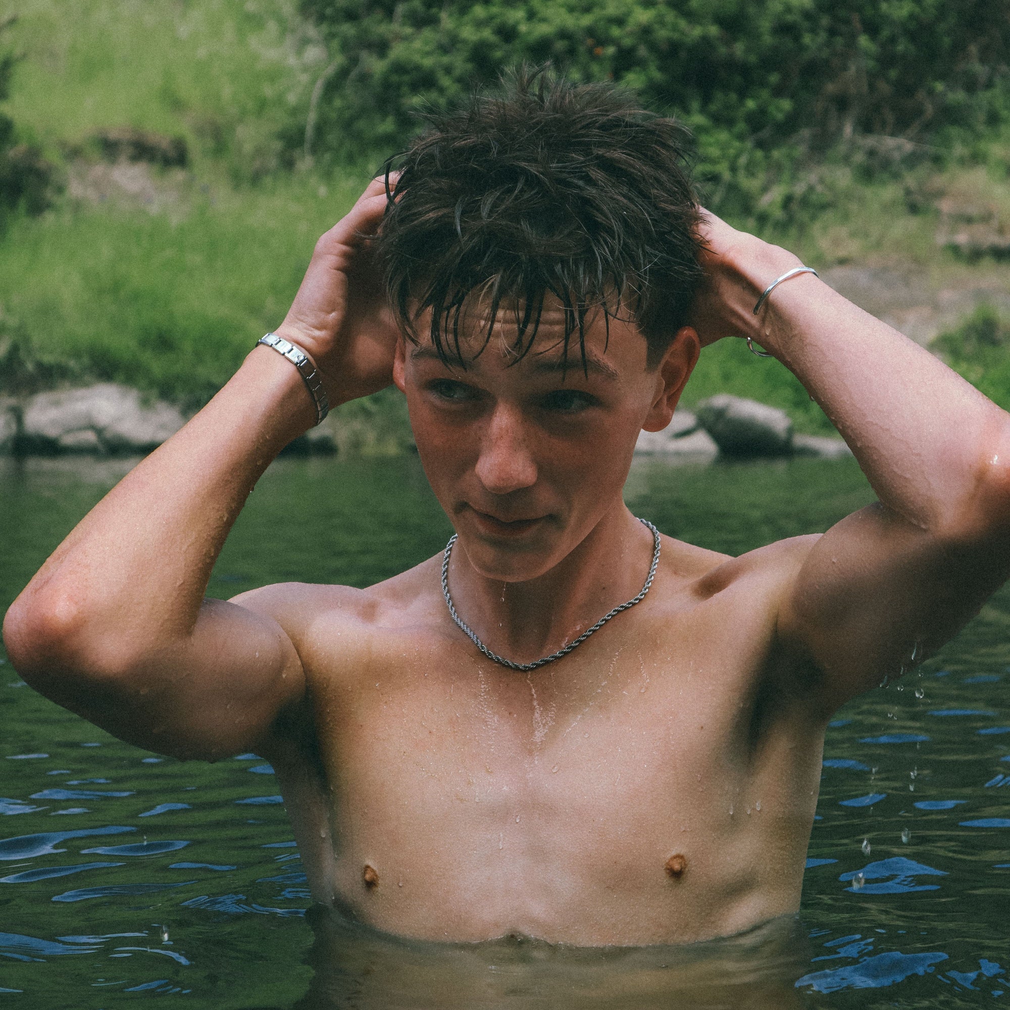 Man with tattoos and wearing jewellery standing in water with a natural background