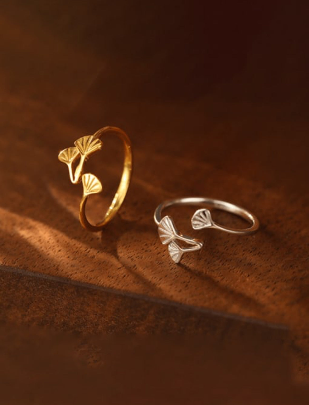 Two rings with a flower plant design, one in gold plating and one in silver, placed on a wooden surface.
