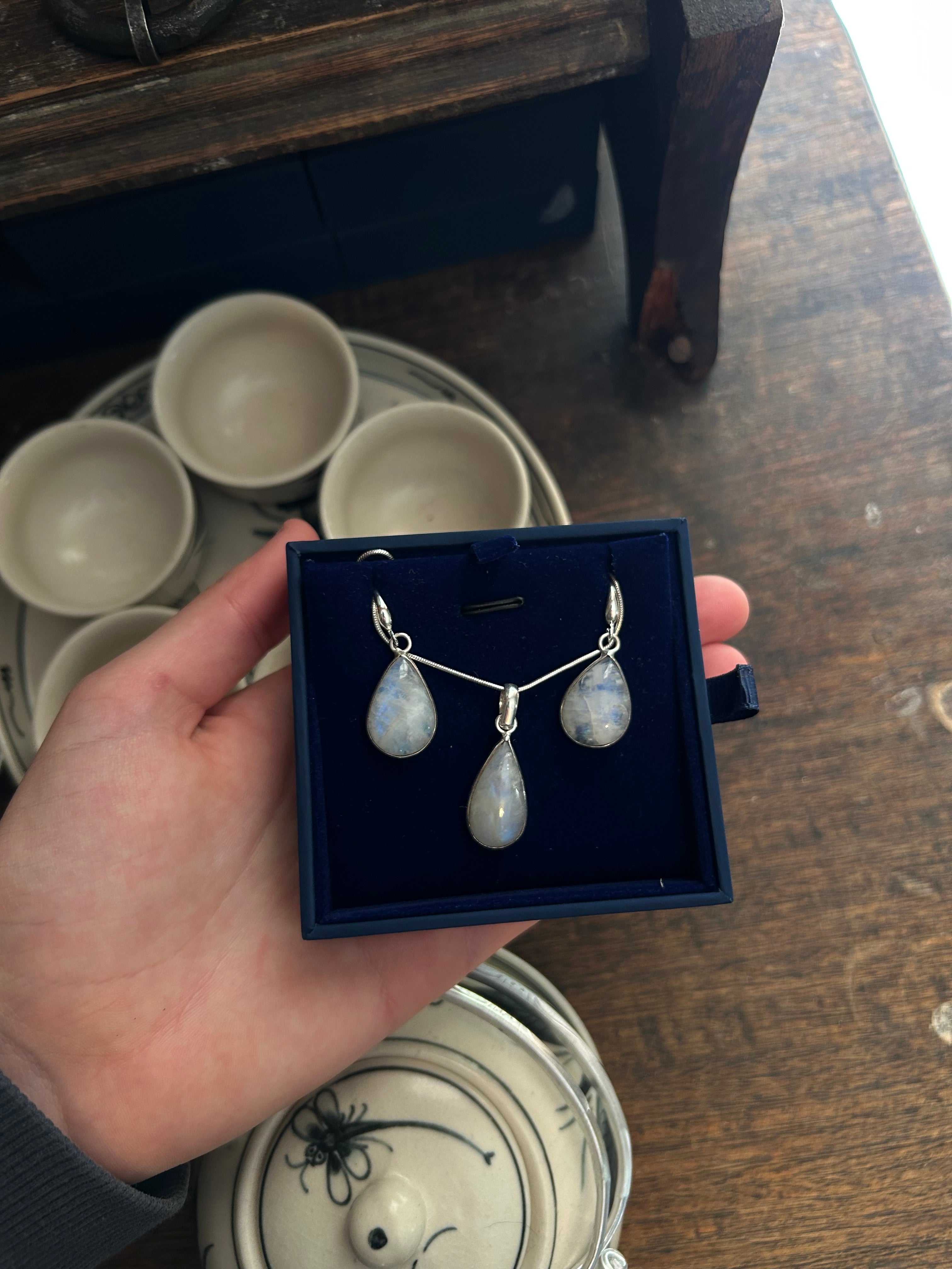 Jewelry set in a box held by a hand on a wooden surface with teacups in the background.