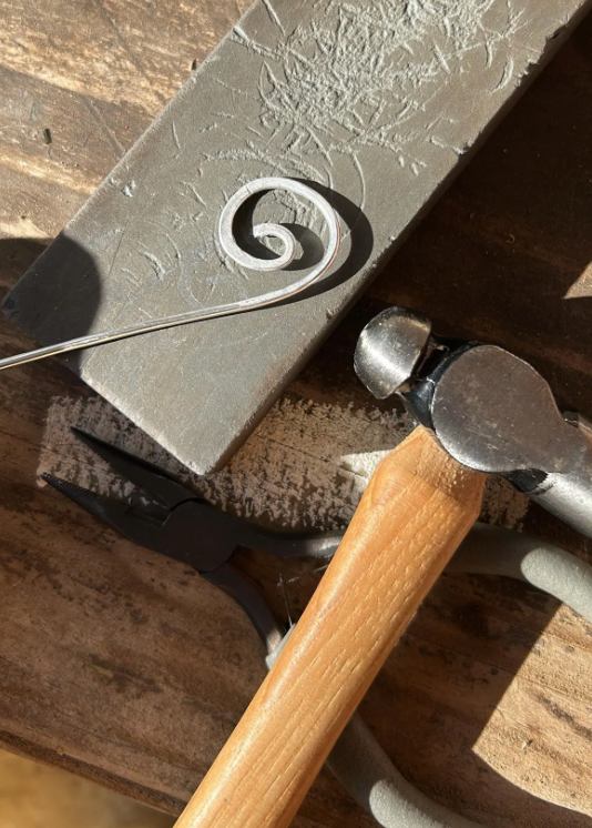 Metalworking tool with a spiral design on metal, surrounded by tools on a wooden surface.