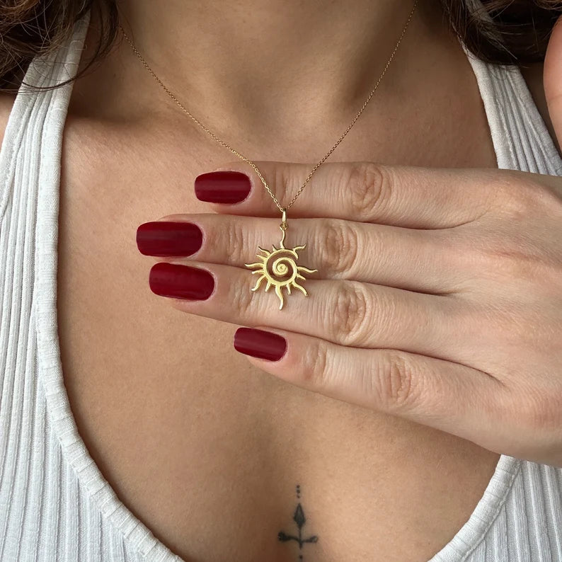 Gold sun-shaped necklace being held by a hand with red nail polish.