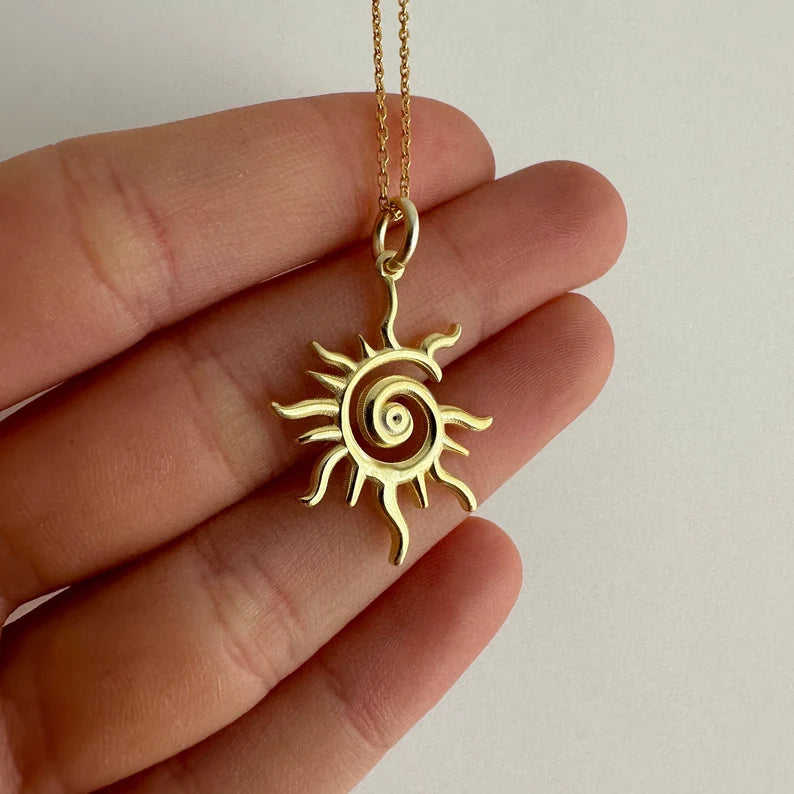 Gold sun-shaped pendant on a chain held between fingers against a neutral background