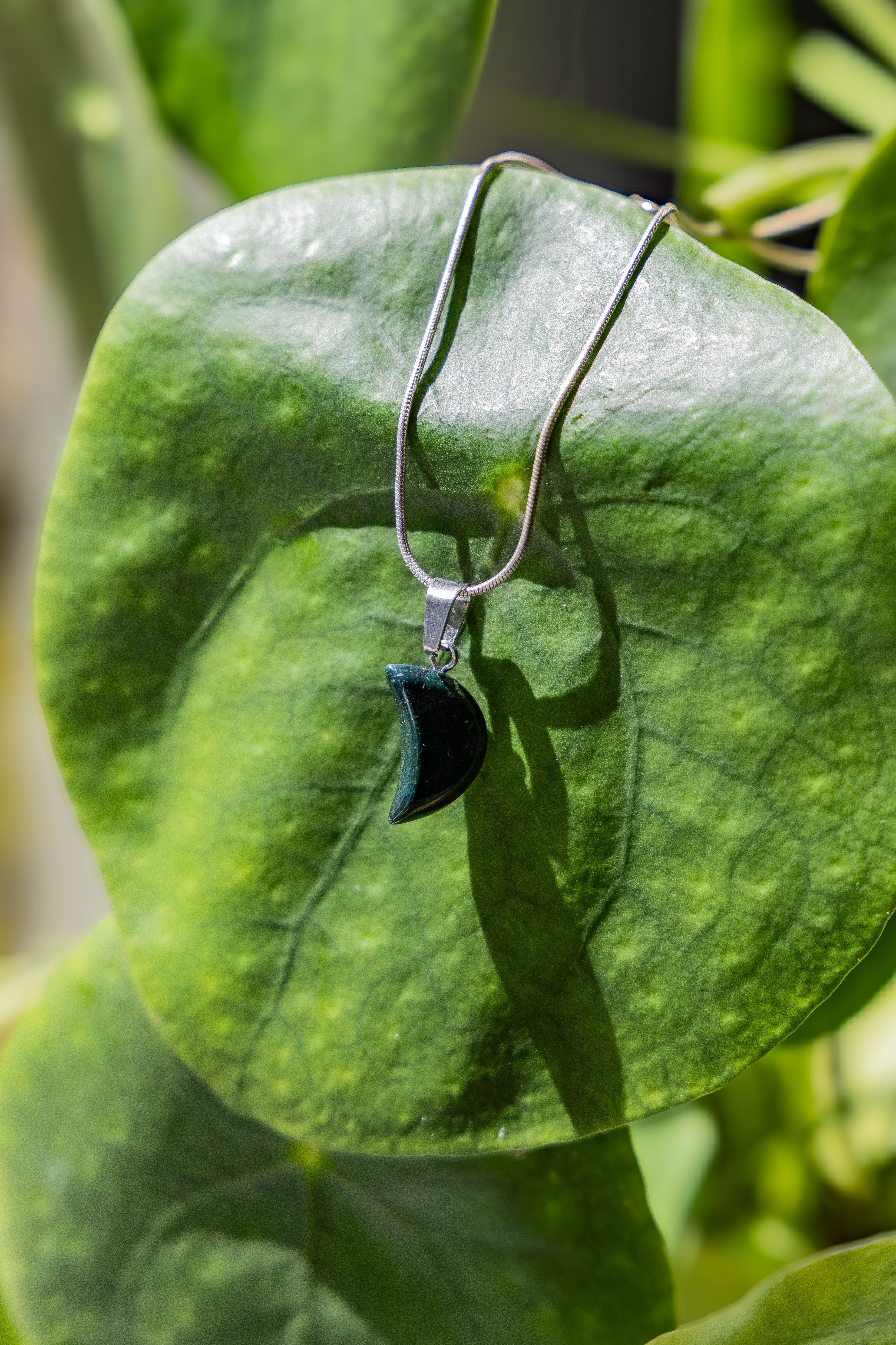 A green moon-shaped pendant necklace displayed on a green leaf.